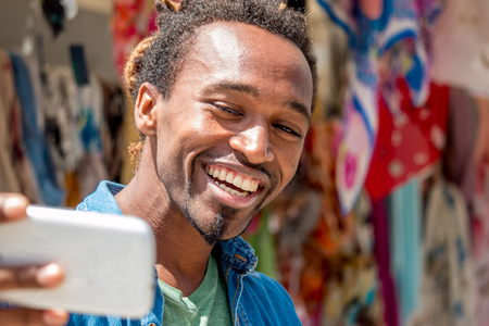 happy african guy is spontaneous, smiling and taking a selfie photo using smartphone out in a colorful market place - cheerful man looking at the phone with a wide toothy smile on a bright sunny dayの写真素材