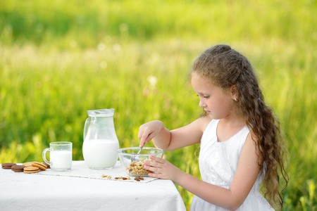 Smiling little girl sitting near the table and having breakfast and drinking milk. Curly cute child sitting outdoor summer gardenの写真素材