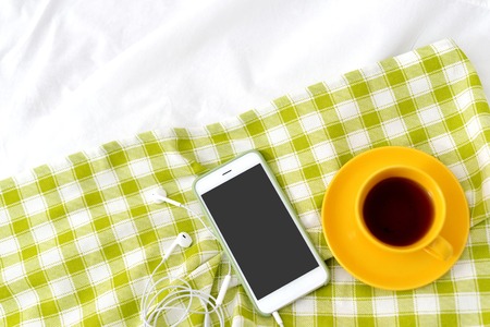 Flat lay phone, yellow cup of tea and flowers on white blanket with green napkinの写真素材