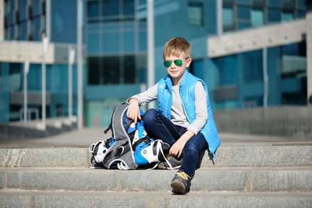 Young boy sitting near grey wall with inline roller skates and all protection at outdoor skate parkの写真素材