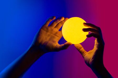 Man holding yellow moon shape illuminated sphere. Surrealistic photo style, contemporary art element for design, posters and banners. Neon blue and purple light.の写真素材