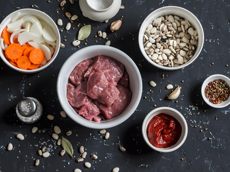 Raw ingredients - beef, beans, onions, carrots, spices, tomato sauce on a dark stone backgroundの写真素材