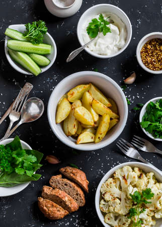 Baked cauliflower and potatoes, fresh cucumbers and herbs, sauces and spices on a dark stone background. Healthy vegetarian lunch or snack in a rustic style. Top viewの写真素材