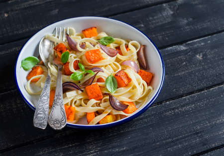 Pasta with baked pumpkin and onion on a white enamel plate on dark wooden surfaceの写真素材