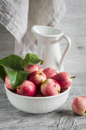 fresh garden apples in a white bowl, vintage enameled pitcher on a light wooden surfaceの写真素材
