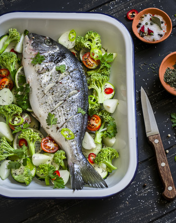 raw ingredients - dorado fish and vegetables - broccoli, zucchini, onions, peppers, lime and spices on a dark wooden surface, cooking baked fish with vegetablesの写真素材
