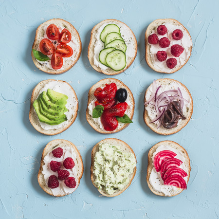 Assortment of sandwiches - sandwiches with cheese, tomatoes, anchovies, roasted peppers, raspberries, avocado, bean pate, cucumber, olives. On a blue background, top view.の写真素材