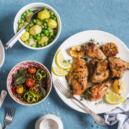 Lunch table. Lemon thyme baked chicken, boiled potatoes with green peas, salad with lentils and tomatoes on a blue background. Top viewの写真素材