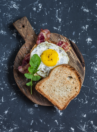 Toast with fried egg, bacon and arugula on the wooden cutting board. Delicious breakfast. On a dark background, top viewの写真素材