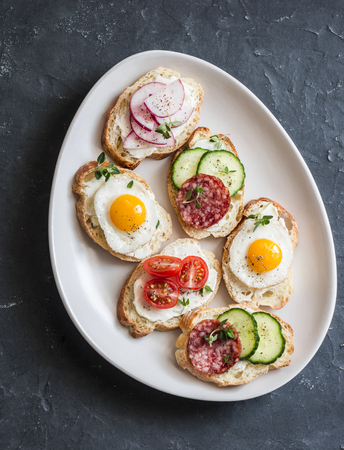 Variety of mini sandwiches with cream cheese, vegetables, quail eggs and salami. Sandwiches with cheese, cucumber, radish, tomatoes, salami, quail eggs on a dark background, top viewの写真素材