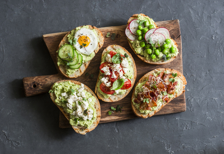 Breakfast avocado sandwich - with crispy bacon, quail egg, tomatoes, goat cheese, green peas, radish, cucumber on rustic cutting board. Healthy snack. On a dark background, top viewの写真素材