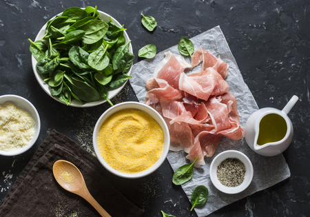 Ingredients for cooking - polenta, spinach and prosciutto. Delicious lunch in a mediterranean style. On a dark background, top view. Flat layの写真素材