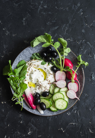 Healthy diet snack in a mediterranean style - greek yogurt, radishes, olives, cucumbers, lettuce on dark background, top view. Flat lay. Copy spaceの写真素材