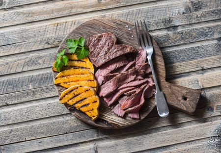 Beef steak and grilled pumpkin on a cutting board on wooden background, top viewの写真素材