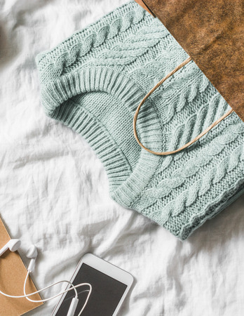 Womens knitted blue sweater in a paper bag and a smartphone with headphones on a light background, top view. Women's clothing shoppingの写真素材