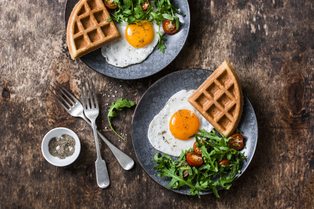 Savory waffles, fried eggs and arugula, cherry tomato salad - delicious healthy breakfast on wooden background, top viewの写真素材