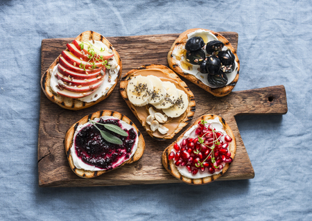 Variety grilled bread dessert small plates sandwiches with cream cheese and apple, pomegranate, jam, grapes, peanut butter, banana, flax seed, chia, nuts on a rustic cutting board on blue background, top view. Flat lay. Delicious breakfast or snackの写真素材