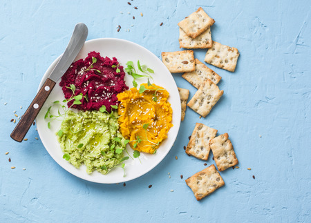 Variations of hummus and crackers. Pumpkin, turmeric, avocado, beetroot hummus with olive oil, sesame seeds and micro greens on a blue background, top viewの写真素材
