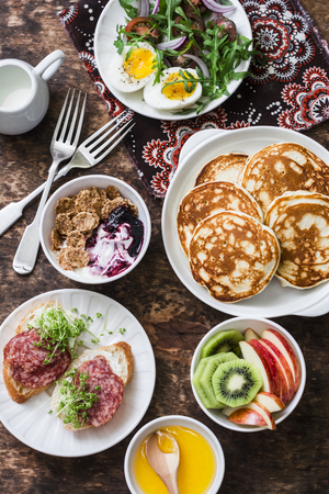 Breakfast table - greek yogurt with whole grain cereals and berry sauce, pancakes, arugula, cherry tomatoes, boiled eggs salad, kiwi, apples fruit, salami and cream cheese sandwiches on a wooden background, top view. Flat layの写真素材