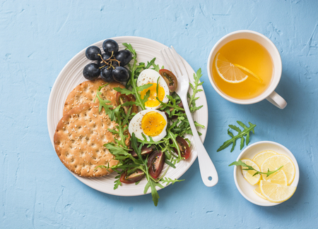 Breakfast plate - cracker, arugula, cherry tomatoes, boiled egg salad and green tea with lemon on blue background, top viewの写真素材