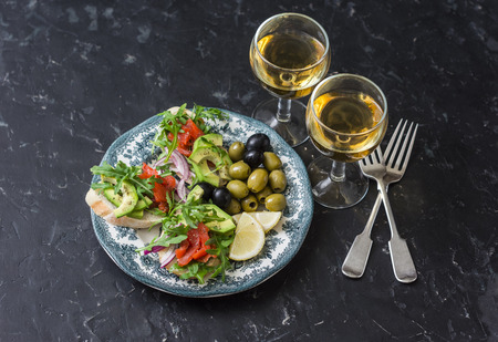 Mediterranean style antipasto and wine. Smoked salmon, avocado, arugula bruschetta, olives and two glasses of white wine on dark backgroundの写真素材