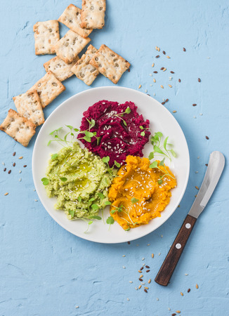 Variations of hummus and crackers. Pumpkin, turmeric, avocado, beetroot hummus with olive oil, sesame seeds and micro greens on a blue background, top viewの写真素材