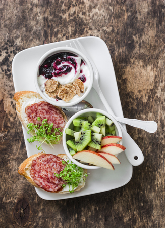 Breakfast or snack plate - salami, cream cheese, micro greens bruschetta, apples, kiwi, yogurt with berries and cereal. Healthy food concept. On wooden background, top viewの写真素材