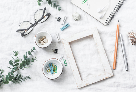 Flat lay still life of homemade crafts work table with accessories - wooden photo frame, flowers, seashells, paper clips, notebook, pencil, decorative clothespins, glasses on a white background, top viewの写真素材