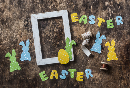 Flat lay Easter background. Empty frame, paper homemade rabbits, ribbon on wooden rustic background, overhead view. Easter decorations home interiorの写真素材