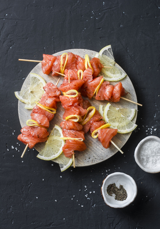 Raw ingredients for cooking salmon skewers on the grill. Raw salmon skewers, lemon, pepper, sea salt on a cutting board on dark background, top viewの写真素材
