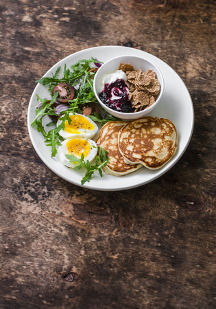 Breakfast plate - boiled egg, arugula, cherry tomatoes salad, greek yogurt with whole grain cereal and blueberries berry sauce, pancakes on wooden background, top view. Healthy food conceptの写真素材