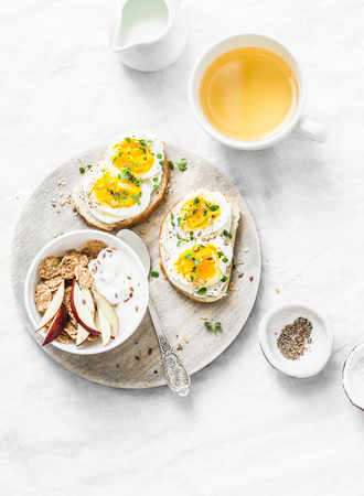 Morning breakfast table inspiration - sandwiches with cream cheese and boiled egg, yogurt with apple and flax seeds, herbal detox tea on light background, top view. Flat layの写真素材