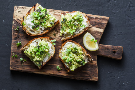 Avocado and ricotta toast with micro greens on a cutting board, on a dark background, top view. Good fats healthy eating concept. Delicious breakfast or snackの写真素材