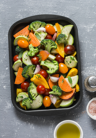 Raw fresh vegetables on a baking sheet. Sweet potato, zucchini, sweet pepper, cherry tomatoes, garlic, broccoli cabbage, olive oil, rose salt - ingredients for healthy vegetarian lunch on grey background, top viewの写真素材