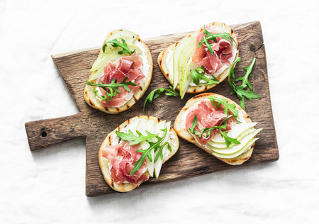 Bruschetta with cream cheese, pear, prosciutto and arugula on wooden chopping board on light background, top viewの写真素材