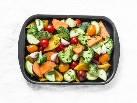 Raw fresh vegetables on a baking sheet. Sweet potato, zucchini, sweet pepper, cherry tomatoes, garlic, broccoli cabbage-ingredients for healthy vegetarian lunch on white background, top viewの写真素材