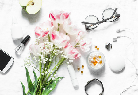 Flat lay women's accessories set - tulips bouquet, cosmetics, glasses, phone, apple, headphones on light background top view. Beauty and fashion conceptの写真素材