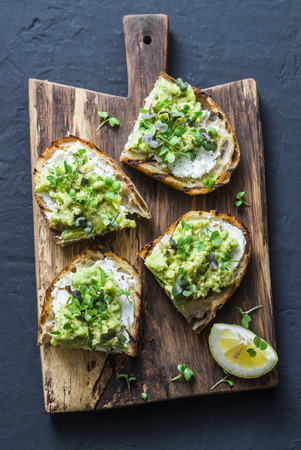 Avocado, cream cheese sandwich with micro greens on a rustic cutting board, top view. Good fats healthy eating concept. Delicious breakfast or snack. On dark backgroundの写真素材
