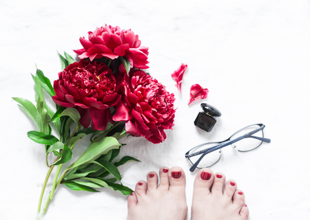Red flowers peonies, beautiful female feet with red pedicure, glasses, perfume on light background, top view. Beauty concept. Womens still life. Soft focus on the flowersの写真素材