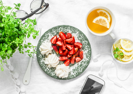 Breakfast or snack table - fresh strawberries, meringue, green tea with lemon, smartphone and headphones. Cozy home rest break still life on a light background, top viewの写真素材