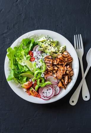 Coconut rice, grilled salmon and vegetables buddha bowl. Healthy lunch - rice, red fish, salad, avocado, radish, tomatoes on a dark background, top viewの写真素材