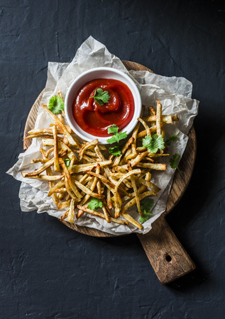 Baked parsnips french fries and homemade ketchup on cutting board - healthy vegetarian snacks on dark background, top viewの写真素材