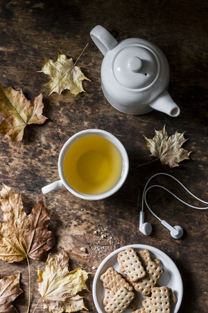 Cozy autumn still life on a wooden background - green tea, whole grain biscuits, headphones, dry maple leaves. Top view, flat layの写真素材