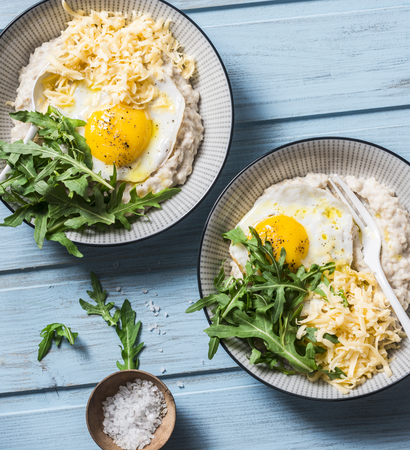 Oatmeal with cheese, fried egg and arugula. Delicious, nutritious breakfast on a blue background, top viewの写真素材