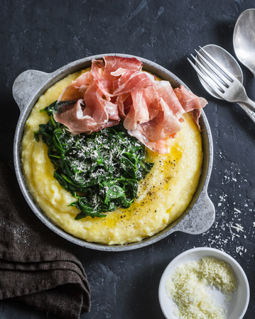 Polenta with butter stewed spinach and prosciutto in a frying pan. Delicious lunch in a mediterranean style. On a dark background, top viewの写真素材