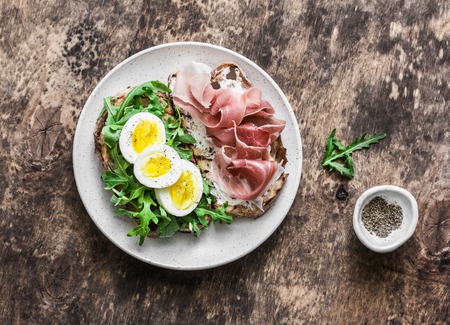 Delicious breakfast, snack, appetizers - sandwiches with prosciutto, egg, arugula and cream cheese on a dark background, top view. Flat layの写真素材