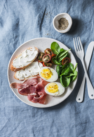 Cream cheese toast, boiled egg, prosciutto, spinach, tomatoes - delicious healthy breakfast or snack on blue background, top viewの写真素材