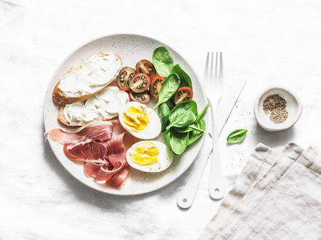 Delicious healthy tapas - cream cheese toast, boiled egg, prosciutto, spinach, tomatoes on light background, top viewの写真素材