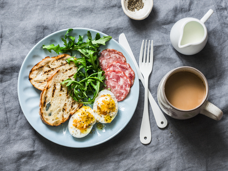 Delicious breakfast or snack - salami sausage, boiled egg, arugula, grilled bread and coffee on a gray background, top viewの写真素材