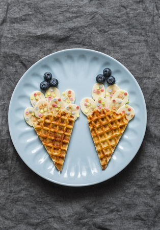 Creative fun baby food. Waffles with banana and blueberries on a gray background, top view. Cheerful children's food. Flat lay, copy spaceの写真素材